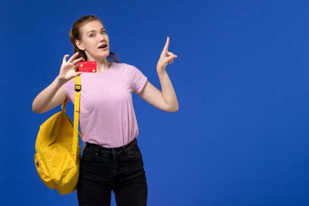 Vista frontal de la mujer joven en camiseta rosa con mochila amarilla sosteniendo una tarjeta roja de plástico en la pared azul