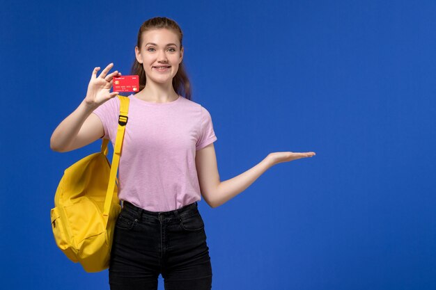 Vista frontal de la mujer joven en camiseta rosa con mochila amarilla sosteniendo una tarjeta roja de plástico sonriendo en la pared azul