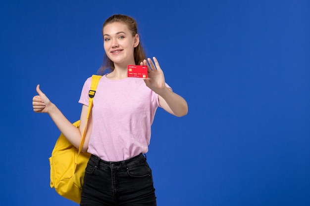 Vista frontal de la mujer joven en camiseta rosa con mochila amarilla sosteniendo una tarjeta roja de plástico sonriendo en la pared azul