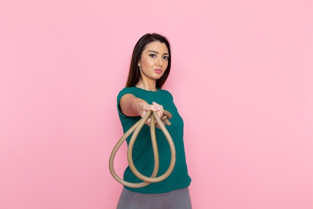 Vista frontal mujer joven en camiseta verde sosteniendo la cuerda para el deporte en la pared rosa cintura deporte ejercicio entrenamientos belleza atleta delgada