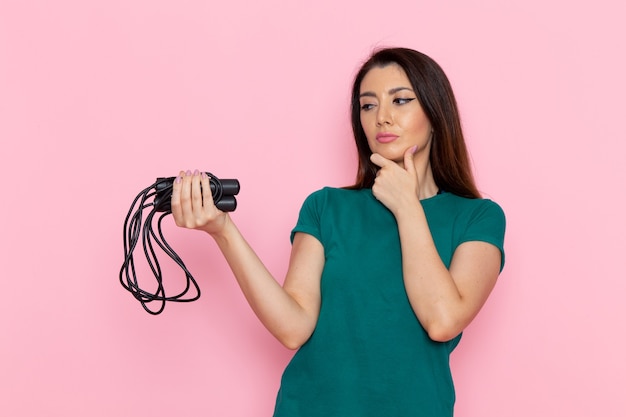 Vista frontal mujer joven en camiseta verde sosteniendo saltar la cuerda y pensando en la pared rosa claro cintura ejercicio entrenamiento belleza delgado deporte femenino