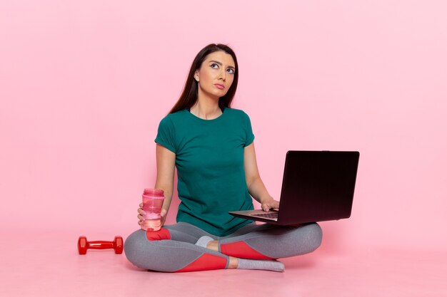 Vista frontal mujer joven en camiseta verde trabajando en la computadora portátil en la pared rosa cintura ejercicio entrenamiento belleza deporte femenino delgado