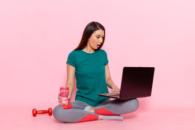 Vista frontal mujer joven en camiseta verde trabajando con su computadora portátil en la pared rosa cintura ejercicio entrenamiento belleza deporte femenino delgado