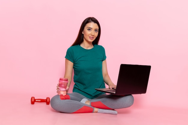 Foto gratuita vista frontal mujer joven en camiseta verde trabajando con su computadora portátil en la pared rosa claro cintura ejercicio entrenamiento belleza delgado deporte femenino