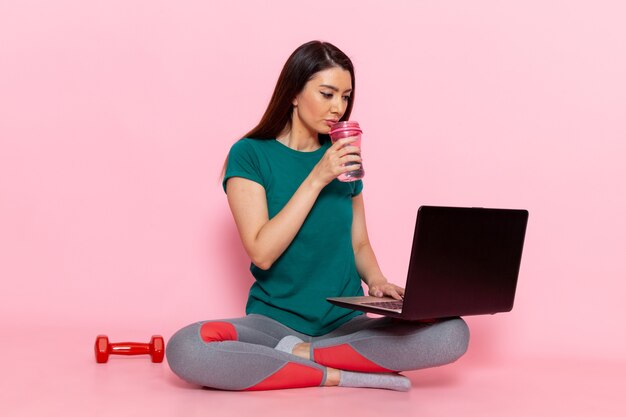 Vista frontal mujer joven en camiseta verde usando su computadora portátil en la pared rosa cintura deporte ejercicio entrenamiento belleza delgado atleta