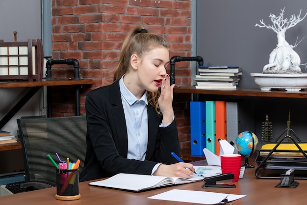 Vista frontal de la mujer joven cansada sentada en una mesa y escribiendo en un documento en la oficina