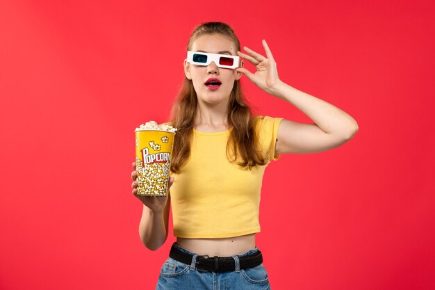 Vista frontal mujer joven en el cine con paquete de palomitas de maíz en gafas de sol d en la pared roja cine cine película divertida femenina