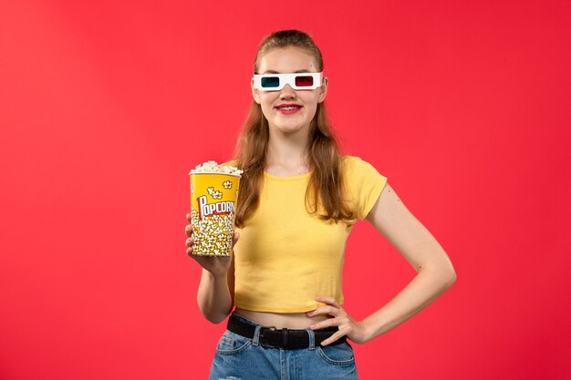 Vista frontal mujer joven en el cine con paquete de palomitas de maíz en gafas de sol d en la pared roja cine cine película divertida femenina