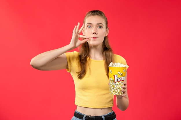 Vista frontal mujer joven en el cine con paquete de palomitas de maíz en la pared de color rojo claro.