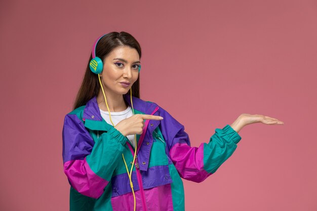 Vista frontal mujer joven en coloridos auriculares de abrigo moderno escuchando música en la pared rosa, ropa de moda de mujer de fotografía en color