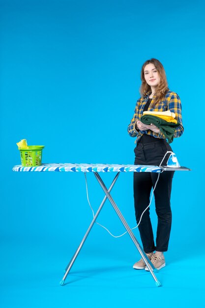 Vista frontal de la mujer joven confiada que sostiene la ropa doblada y de pie detrás de la tabla de planchar en el lavadero
