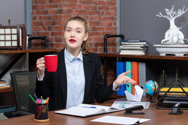 Foto gratuita vista frontal de la mujer joven confundida sentada en una mesa y sosteniendo una taza roja en la oficina