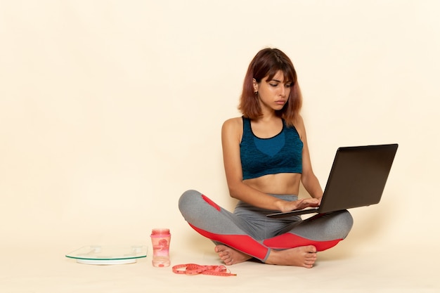 Vista frontal de la mujer joven con cuerpo en forma en camisa azul con laptop en la pared blanca clara