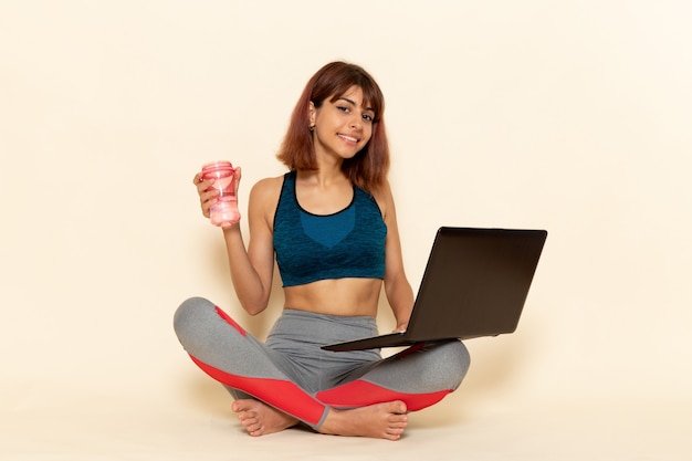 Vista frontal de la mujer joven con cuerpo en forma en camisa azul con laptop en la pared blanca clara