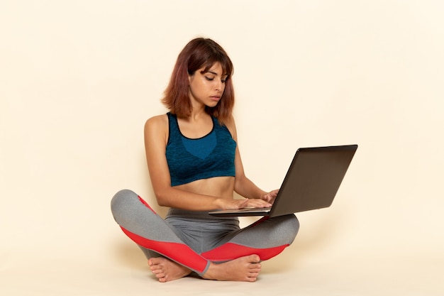 Vista frontal de la mujer joven con cuerpo en forma en camisa azul con ordenador portátil en la pared blanca
