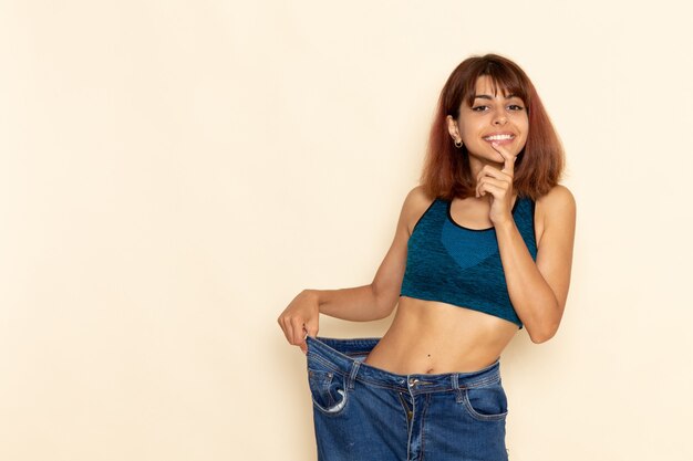 Vista frontal de la mujer joven con cuerpo en forma en camisa azul sonriendo en la pared de color blanco claro