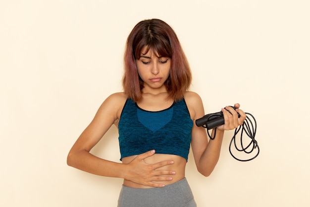 Foto gratuita vista frontal de la mujer joven con cuerpo en forma en camisa azul sosteniendo cuerdas para saltar en la pared blanca clara