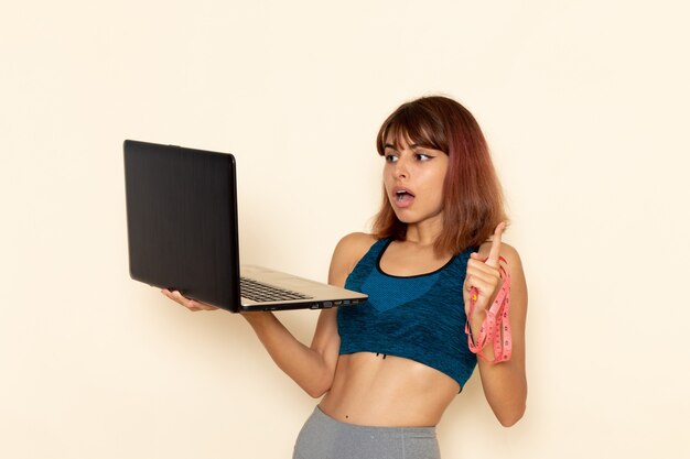 Vista frontal de la mujer joven con cuerpo en forma en camisa azul usando su computadora portátil en la pared blanca clara