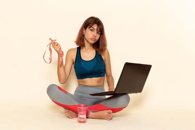 Vista frontal de la mujer joven con cuerpo en forma en camisa azul usando su computadora portátil en la pared blanca clara