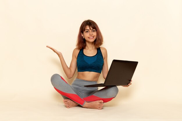 Vista frontal de la mujer joven con cuerpo en forma en camisa azul usando su computadora portátil en la pared blanca