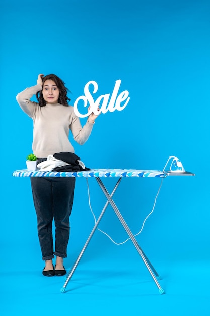 Vista frontal de la mujer joven decepcionada de pie detrás de la tabla de planchar y mostrando el icono de venta en la pared azul