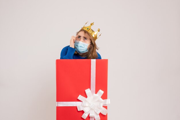 Vista frontal de la mujer joven dentro de la caja de regalo en máscara en la pared blanca