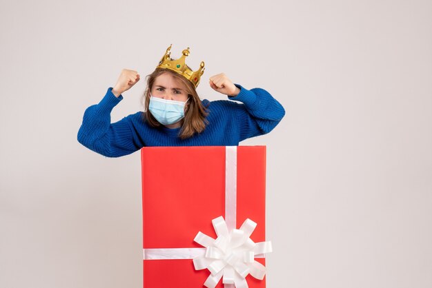 Vista frontal de la mujer joven dentro de la caja de regalo en máscara en la pared blanca