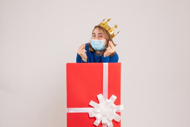 Vista frontal de la mujer joven dentro de la caja de regalo en máscara en la pared blanca