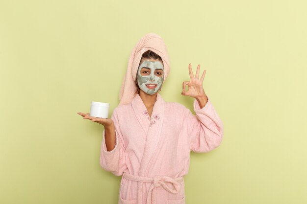 Vista frontal mujer joven después de la ducha en albornoz rosa sosteniendo crema y sonriendo sobre una superficie verde