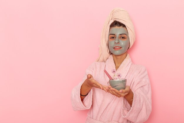 Vista frontal mujer joven después de la ducha en bata de baño rosa aplicando máscara sobre superficie rosa claro