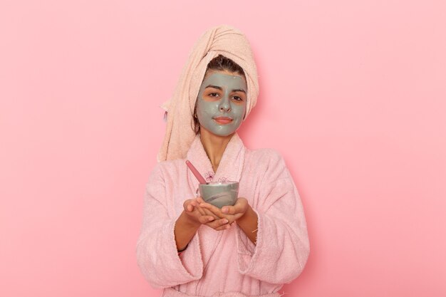 Vista frontal mujer joven después de la ducha en bata de baño rosa aplicando máscara sobre superficie rosa claro