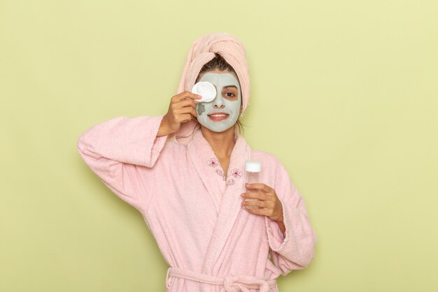 Vista frontal mujer joven después de la ducha en bata de baño rosa quitando su máscara sobre una superficie verde