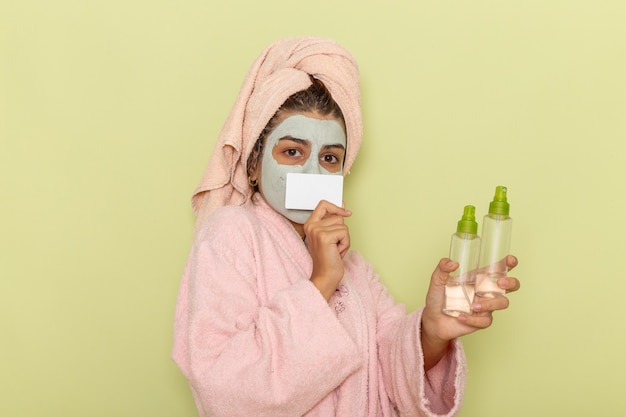 Vista frontal mujer joven después de la ducha en bata de baño rosa sosteniendo aerosoles y tarjeta en superficie verde claro