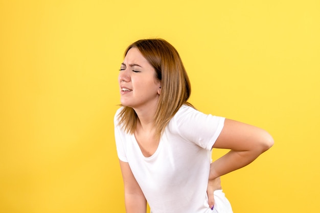 Vista frontal de la mujer joven con dolor de espalda en la pared amarilla