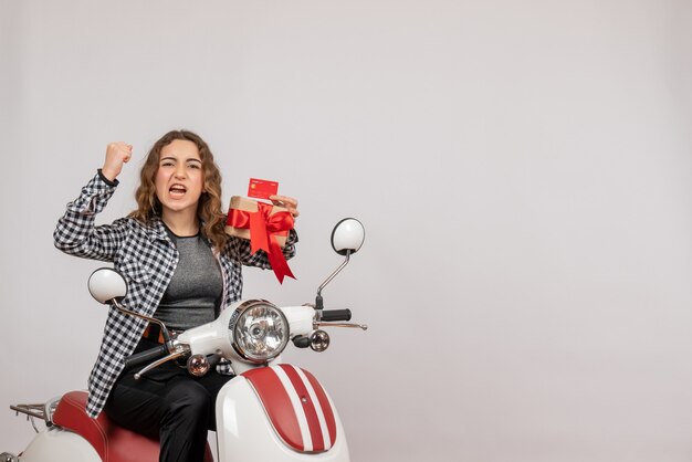 Vista frontal de la mujer joven enojada en ciclomotor con regalo en pared gris