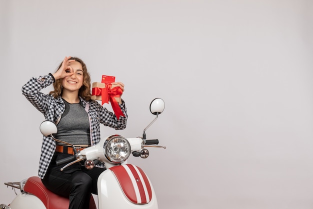 Vista frontal de la mujer joven feliz en ciclomotor sosteniendo binoculares de mano haciendo regalos en la pared gris