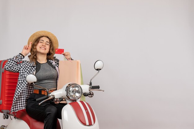 Vista frontal de la mujer joven feliz en ciclomotor sosteniendo la tarjeta en la pared gris