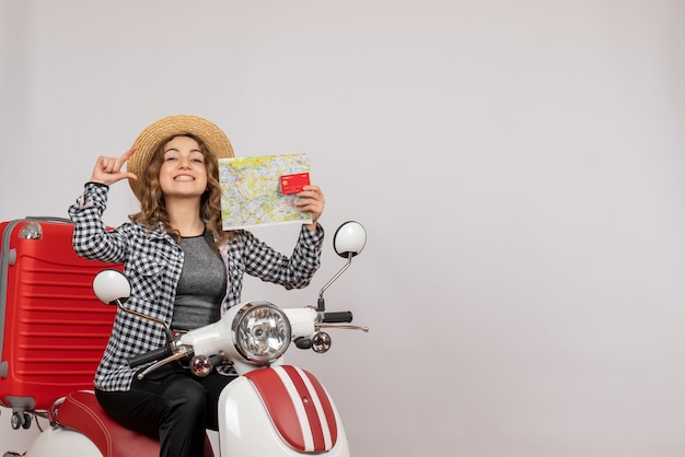 Vista frontal de la mujer joven feliz en ciclomotor con tarjeta y mapa en pared gris