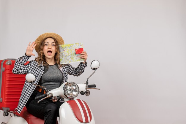 Vista frontal de la mujer joven feliz en ciclomotor con tarjeta y mapa en pared gris