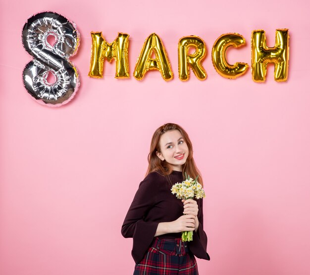 Vista frontal mujer joven con flores en sus manos y decoración de marzo sobre fondo rosa fiesta día de la mujer marzo igualdad matrimonio pasión presente