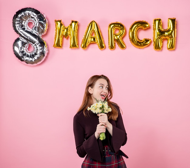 Vista frontal mujer joven con flores en sus manos y decoración de marzo sobre fondo rosa presente día de la mujer marzo matrimonio pasión fiesta igualdad sensual