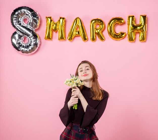 Vista frontal mujer joven con flores en sus manos y decoración de marzo sobre fondo rosa presente día de la mujer marzo pasión fiesta igualdad sensual