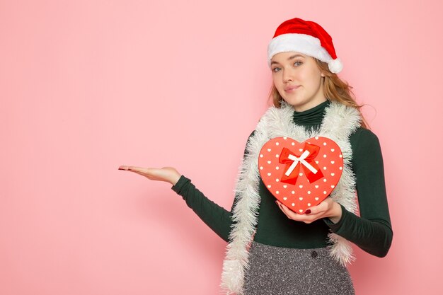 Vista frontal de la mujer joven en gorra roja con presente en forma de corazón en la pared rosa