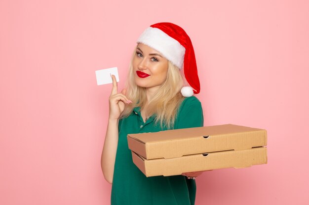 Vista frontal de la mujer joven con gorra roja sosteniendo cajas de pizza y tarjeta bancaria en la pared rosa