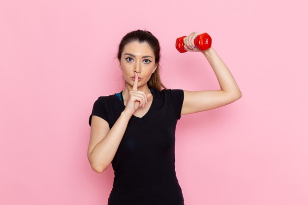 Vista frontal mujer joven haciendo deporte y sosteniendo pesas en la pared rosa atleta deporte ejercicio salud entrenamientos