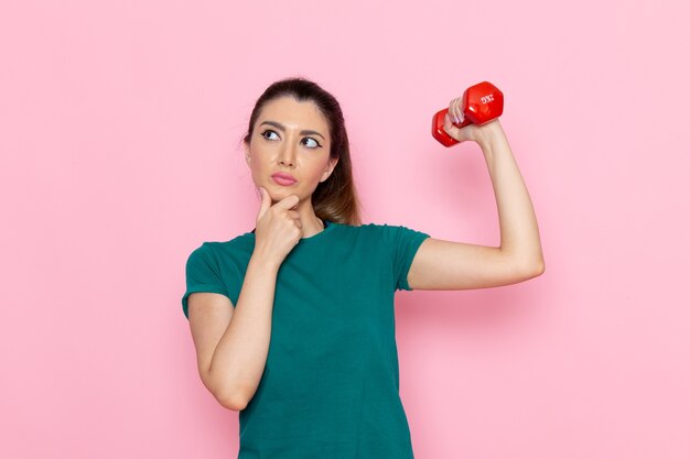 Vista frontal mujer joven haciendo deporte y sosteniendo pesas en la pared rosa atleta deporte ejercicio salud entrenamientos