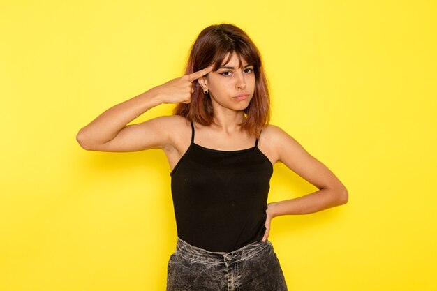 Vista frontal de la mujer joven en jeans gris camisa negra posando en la pared amarilla