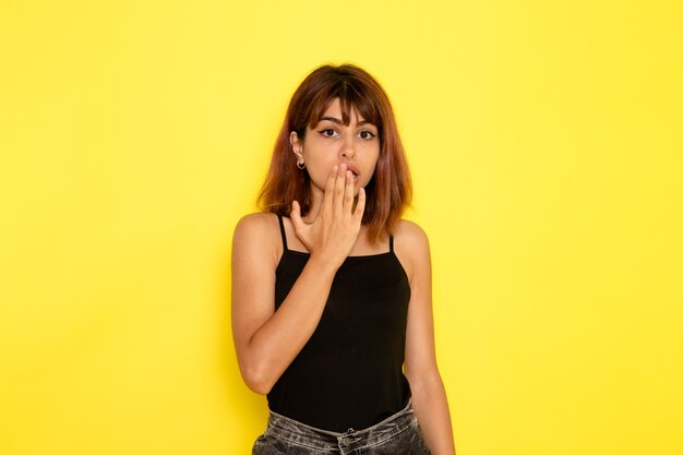 Vista frontal de la mujer joven en jeans gris camisa negra posando en la pared amarilla