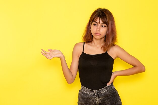 Vista frontal de la mujer joven en jeans gris camisa negra posando en la pared amarilla