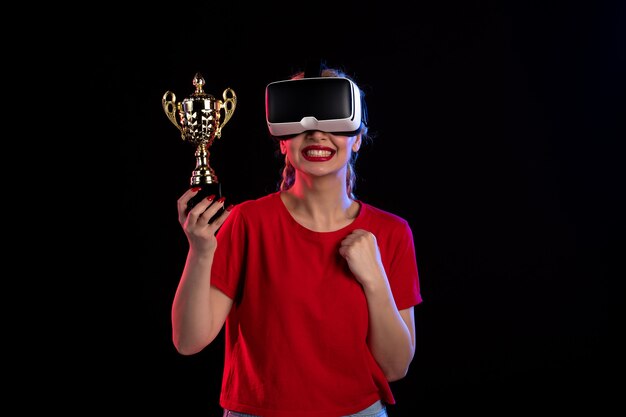 Vista frontal de la mujer joven jugando vr y ganando la copa en la pared oscura